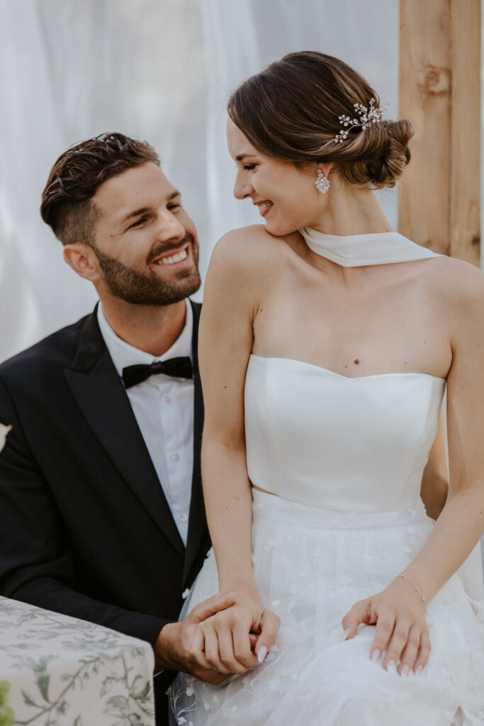 Brautpaar lächelnd, die Braut auf dem Schoß des Bräutigams auf ihrer internationalen Hochzeit