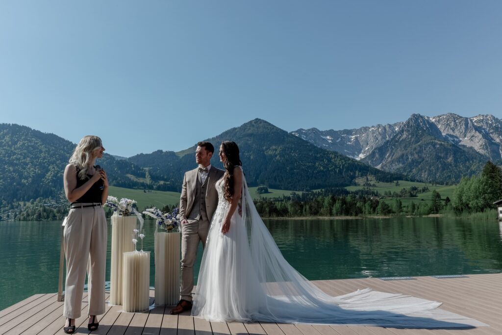 Trauredner Evylin steht bei einer freien Trauung auf einem Steg zusammen mit dem Brautpaar, im Hintergrund hat die Hochzeit im Ausland eine Berg- und Seekulisse