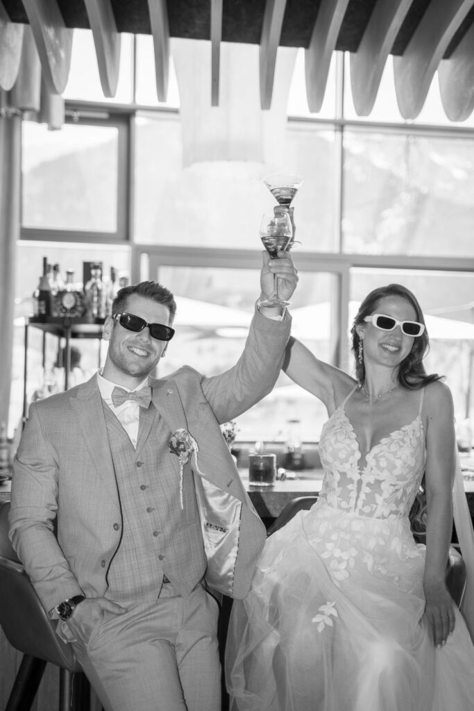 Hochzeitspaar mit Sonnenbrillen hebt auf ihrer Auslandshochzeit glücklich ihre Drinks in die Höhe